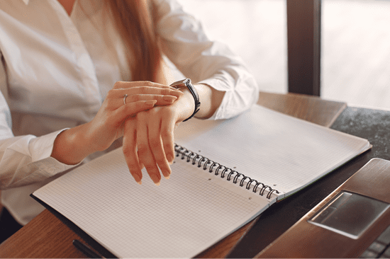 Photo of a woman checking her watch for the time while a scheduling book lies open on the desk in front of her
