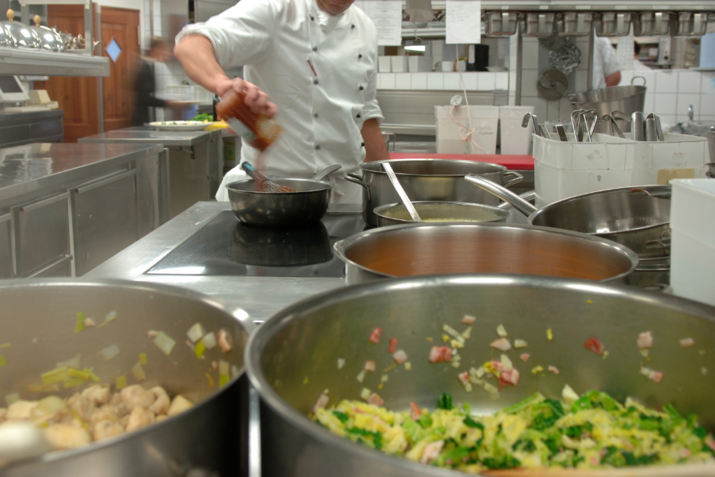 A chef adding salsa to a pot with many other cooking pots in the foreground.