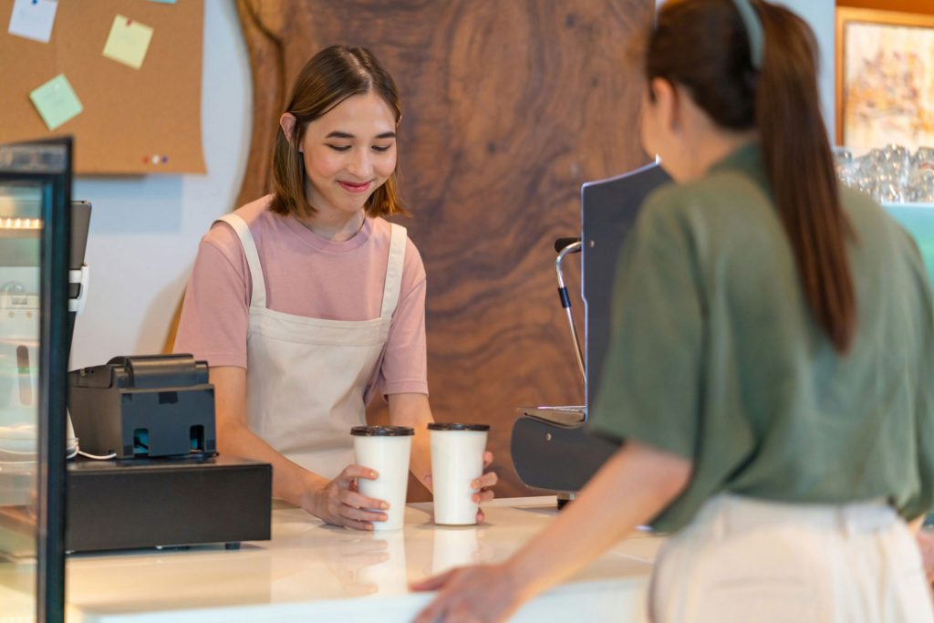 Why The Best Teams Are Using Gamification To Stay Ahead 1 A smiling barista hands two coffee cups to a customer at the counter, illustrating how gamification can motivate employees and improve upsell performance in a café setting.